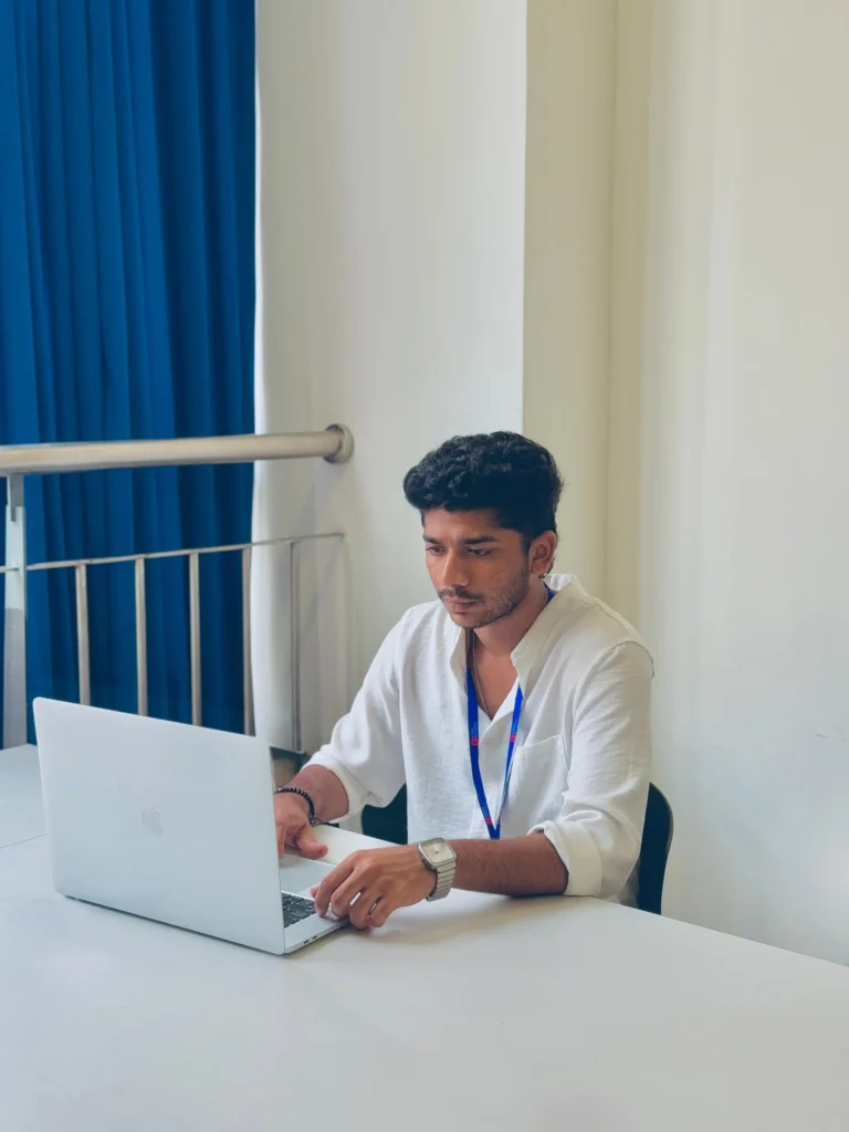 A young man, who is the digital marketing specialist in calicut, is intently working on a silver laptop at a clean white desk, wearing a white linen shirt and a blue lanyard.