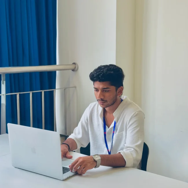 A young man, who is the digital marketing specialist in calicut, is intently working on a silver laptop at a clean white desk, wearing a white linen shirt and a blue lanyard.